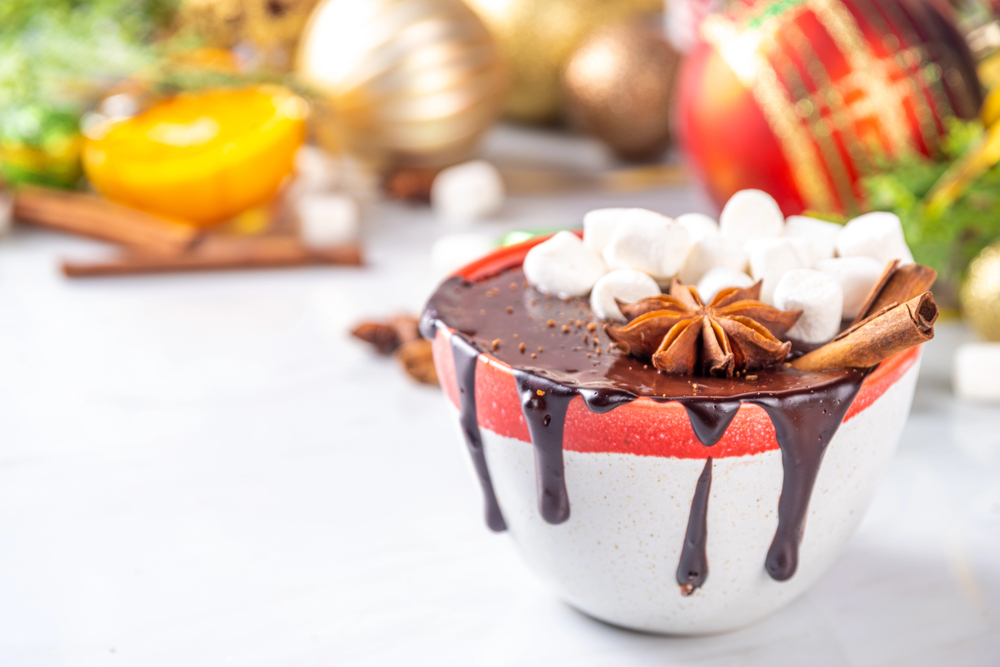 hot chocolate slightly overflowing from cup, with star anise, a cinnamon stick and marshmallow. soft focus festive background